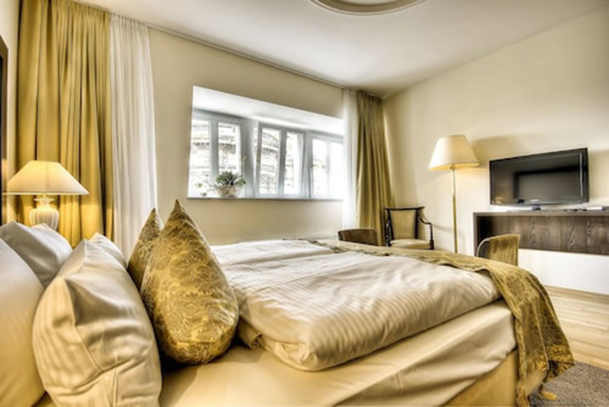 Hotel Suitess Δρέσδη Εξωτερικό φωτογραφία The photo shows a cozy and modern hotel room or bedroom. There is a neatly made bed with soft white linens and decorative pillows. To the side, there is a small lamp providing warm light. The room features large curtains, with one set slightly drawn 