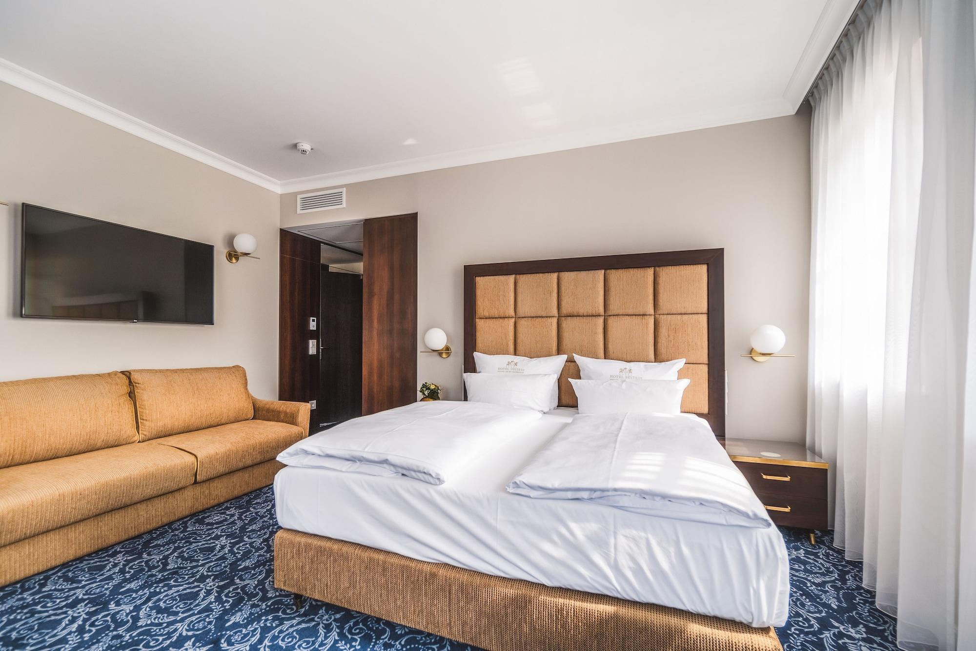 Hotel Suitess Δρέσδη Εξωτερικό φωτογραφία The photo shows a modern hotel room. It features a large bed with white bedding and plush pillows. There is a brown, tufted headboard behind the bed. To the left, there's a comfortable-looking beige sofa, and a flat-screen TV mounted on the wall oppo