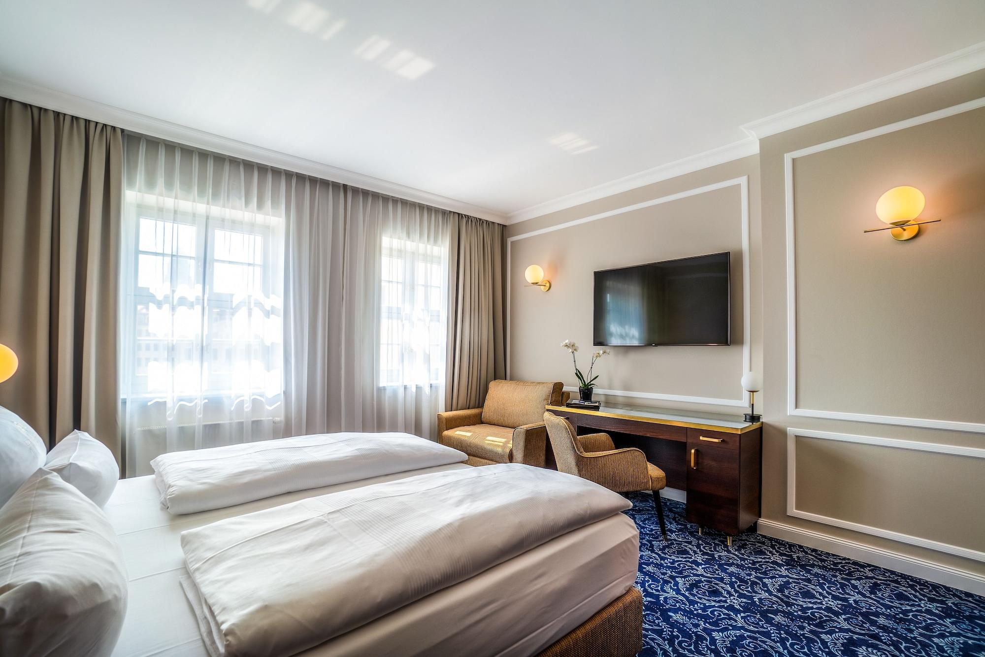 Hotel Suitess Δρέσδη Εξωτερικό φωτογραφία The photo shows a well-furnished hotel room. It features two beds with white bedding positioned side by side. There are large windows covered with light curtains that let in natural light. On one side of the room, there is a comfortable armchair and 