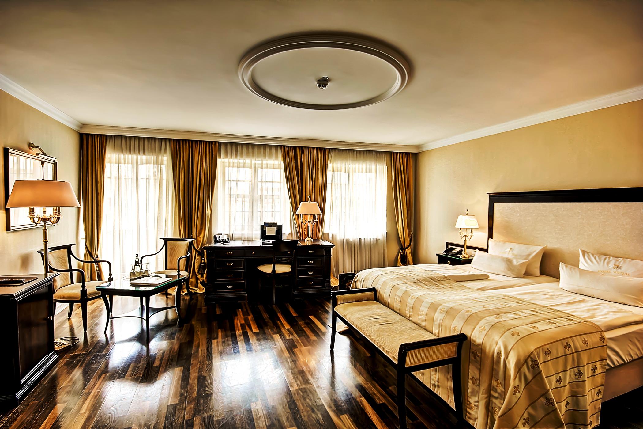 Hotel Suitess Δρέσδη Εξωτερικό φωτογραφία The image shows a stylish hotel room with a warm, elegant aesthetic. It features two beds pushed together, adorned with a light-colored patterned bedspread. Along one wall, there are large windows with sheer curtains that allow natural light to enter