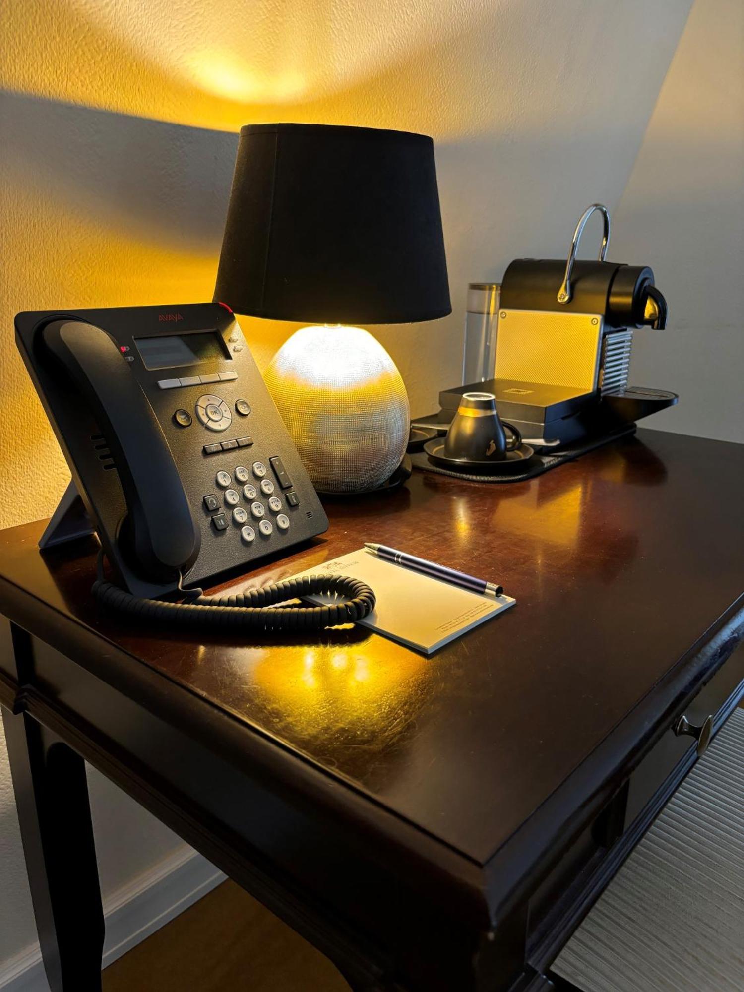 Hotel Suitess Δρέσδη Εξωτερικό φωτογραφία The photo shows a small workspace or side table with several items. There is a black telephone with a keypad on the left side. Next to it is a lamp with a rounded base emitting a warm glow. To the right, there is a sleek coffee machine, likely a Nesp