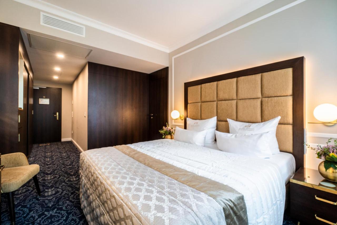 Hotel Suitess Δρέσδη Εξωτερικό φωτογραφία The photo shows a hotel room with a modern design. It features a large bed with a plush headboard, dressed in light-colored bedding. There are two pillows arranged neatly on the bed. To the left, there's a chair and a small decorative table with flow