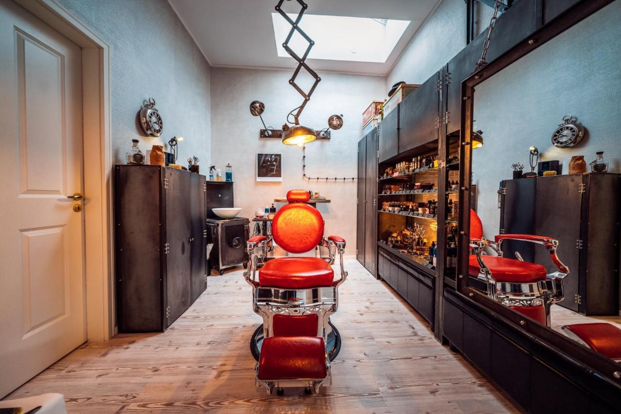 Hotel Suitess Δρέσδη Εξωτερικό φωτογραφία The photo shows a stylish barbershop interior. In the center, there is a classic red barber chair, surrounded by wooden flooring. To the left, there are black cabinets and shelves filled with various grooming products and tools. The room features a l